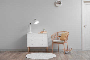 Chest of drawers with glowing lamp, flowerpot, books and wicker chair near light wall