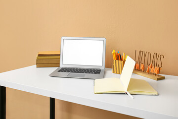 Modern laptop, opened notebook and stationery holder on table near color wall