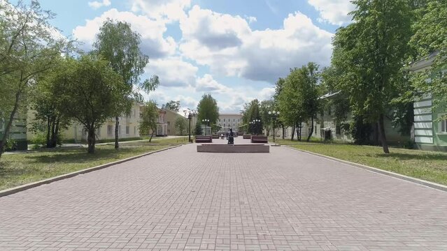 Drone view of a girls with a stroller walks along a beautiful alley in a provincial city with low buildings. Poplar fluff flies. Alley with sculptures, lanterns, benches, flower beds and trees. 
