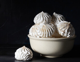 Small white meringues in white ceramic bowl and one meringue nearby on a dark wooden rustic table