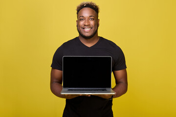 Joyful guy African nationality holds laptop with two hands, unfolded to audience screen. Dark screen of modern laptop shows dark-skinned man with smile on his face in his hands on yellow background