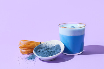 Glass of blue matcha tea, chasen and bowl with powder on purple background