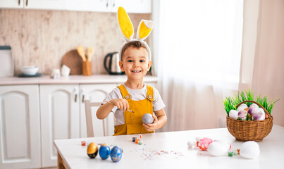 a handsome little boy of European appearance laughs and draws on Easter eggs at the table, on his head with yellow rabbit ears.