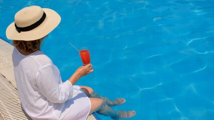 blonde woman in straw hat relaxes and drinks orange cocktail in the swimming pool on a sunny summer day. Vacation concept, all inclusive