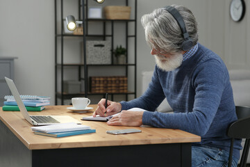 Middle aged man with laptop, notebook and headphones learning at table indoors
