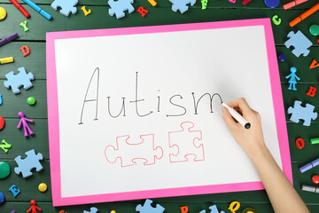 MYKOLAIV, UKRAINE - DECEMBER 30, 2021: Woman writing word Autism on white board at green wooden table with toys, top view