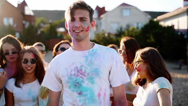 Handsome man stained in colored powder punches cheast and shows peace sign with his fingers at Holi festival. Pretty women jump, dance. Hindu holiday. End of lockdown, covid pandemic, restrictions