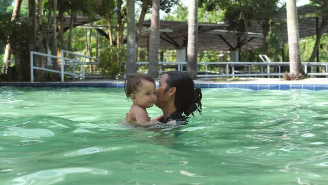 Lovely mother with baby in the pool swim together. Water is good for your baby's health. The kid swims with pleasure in the water and hits the water with his boat creating splashes on the water