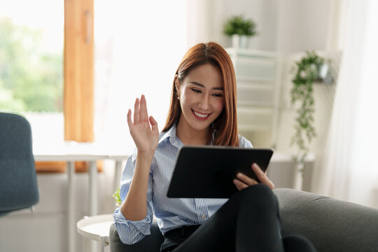 Image of happy asian woman smiling and waving hand at digital tablet, while speaking or chatting on video call at home