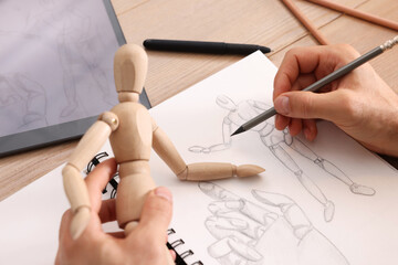 Man drawing mannequin in sketchbook with pencil at wooden table, closeup