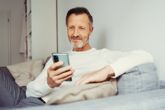 older man sitting at home on sofa and looking at his cell phone
