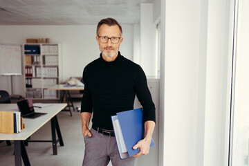 older businessman with glasses holds a folder and looks into the camera