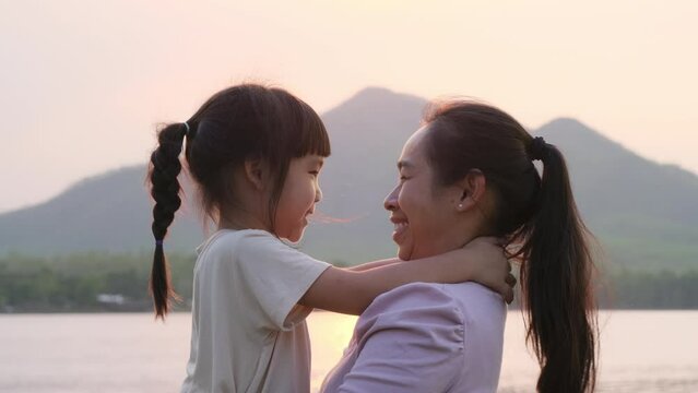 Mother holding a cute little daughter walking by the lake at sunset. Excited kid girl laughs playing with her mother outdoors. Mother hugging her daughter.