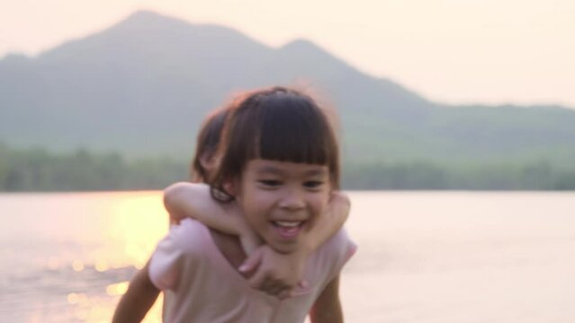 Asian Little girl riding on her elder sister's back walking by the lake at sunset. Happy sisters play together happily outdoors.