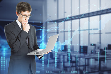 Attractive thoughtful young european businessman with laptop standing in blurry office interior with creative glowing business chart. Growth, sales and market concept. Double exposure.