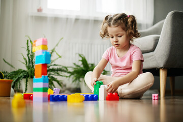 Obraz na płótnie Canvas A little girl is sitting on the floor in the playroom and playing with colorful cubes, a constructor.
