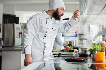 Master chef tasting gourmet dish while cooking food for dinner service at fine dining restaurant. Skilled cook checking meal taste after added fresh organic vegetables and ingredients in kitchen.