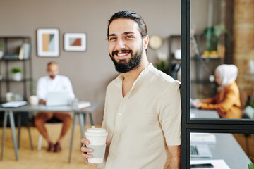 Young cheerful chief executive officer or business leader holding drink while standing in large modern open space office