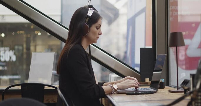 Side view of caucasian woman working in a coffee shop. Woman writing about new project on digital laptop listening music with her headphones. Remote working jobs. Footage made through the window