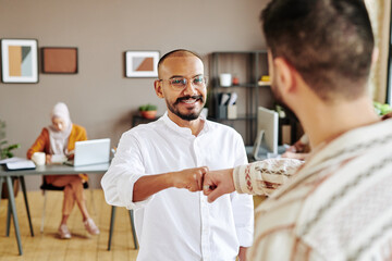 Obraz premium Happy young businessman in casualwear greeting his colleague with punch bump and looking at him while standing in office