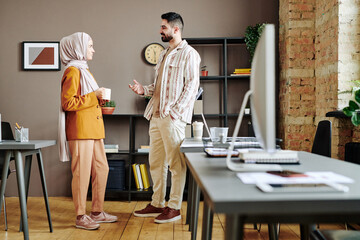 Two young contemporary employees in casual clothes communicating at coffee break while standing in spacious modern openspace office - obrazy, fototapety, plakaty