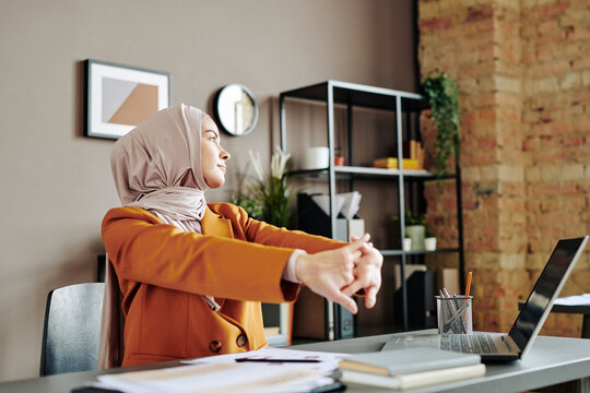 Young contemporary Muslim businesswoman doing stretch exercise while sitting by workplace and looking aside in office