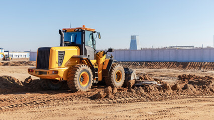 Fototapeta premium Bulldozer or loader moves the earth at the construction site against the blue sky. An earthmoving machine is leveling the site. Construction heavy equipment for earthworks.