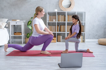Young mother and little daughter exercising online on fitness mat at home with laptop