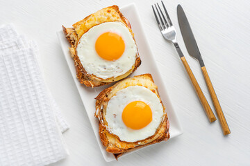 Top view of Croque Madame French hot sandwiches made with ham and cheese with fried egg on top baked or fried served on plate as quick snack for breakfast on white wooden table with towel and fork