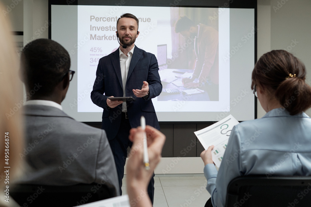 Poster contemporary young male economist in formalwear speaking in microphone by his mouth during presentat