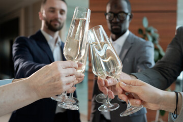 Hands of interracial entrepreneurs with flutes of champagne toasting at buffet while celebrating successful finishing of conference