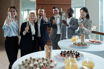 Group of cheerful business team in formalwear toasting at buffet in restaurant while standing by...