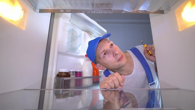 The refrigerator repairman opens the door and examines the inside of the refrigerator with a screwdriver in his hand. A young Caucasian man in a blue jumpsuit or uniform. Refrigerator repair.
