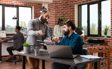 Millennial entrepreneur men discussing marketing strategy analyzing company graph while working at business project in brick wall startup office. Multi-ethnic team brainstorming ideas