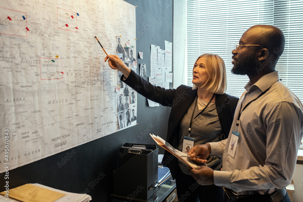 Poster two fbi agents looking at map on board while mature female pointing at it and making suggestions abo