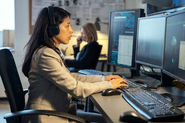 Side view of young Hispanic female operator of surveillance system sitting by desk in front of...