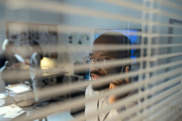 Young serious agent of FBI or secret service speaking on mobile phone while standing by window with...