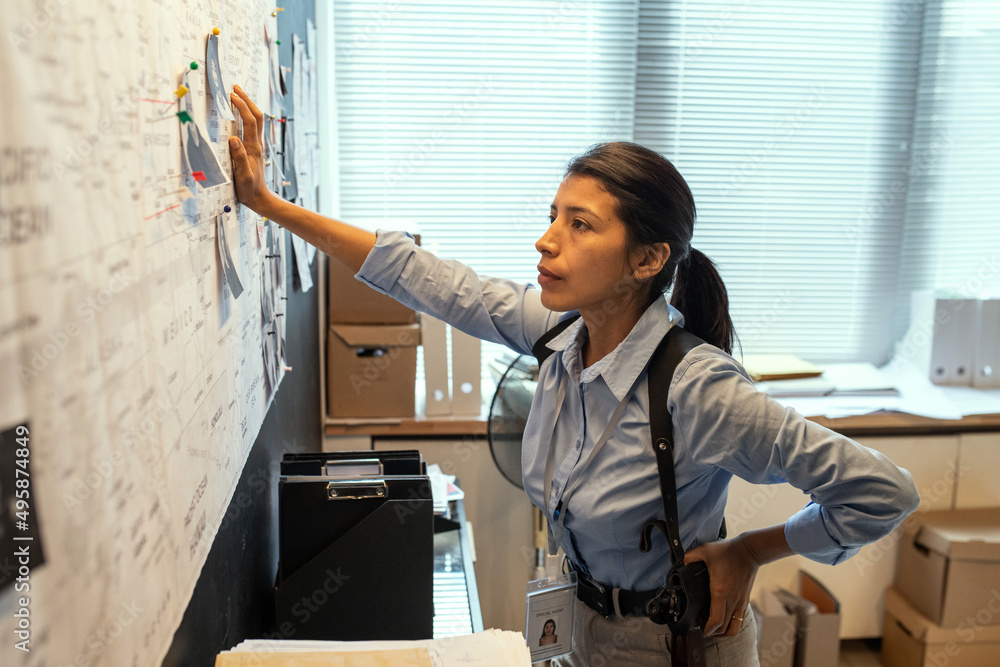 Poster young serious fbi agent in uniform looking at map with pinned photos of criminals in office while th