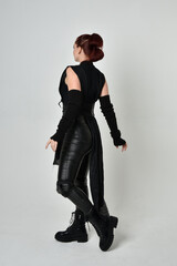 Full length portrait of pretty red haired female model wearing black futuristic scifi leather costume. Dynamic standing poses with gestural hands, facing backwards away from  on a white studio backgro