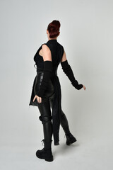 Full length portrait of pretty red haired female model wearing black futuristic scifi leather costume. Dynamic standing poses with gestural hands, facing backwards away from  on a white studio backgro