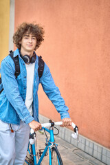 Young curly-haired student with headphones walking to college holding bicycle with hands looking at...