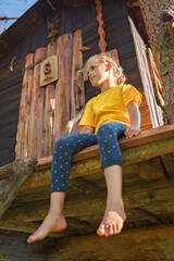 Pretty girl sitting upstairs in beautiful creative handmade treehouse in backyard, summer activity, cottagecore, happy summertime in countryside, ecological outdoor playground, lower angle