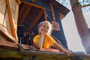 Happy pretty girl looking down from the beautiful creative handmade treehouse in backyard, summer...