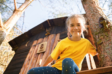 Fototapeta na wymiar Happy pretty girl looking down from the beautiful creative handmade treehouse in backyard, summer activity, cottagecore, happy summertime in countryside, ecological outdoor playground, lower angle