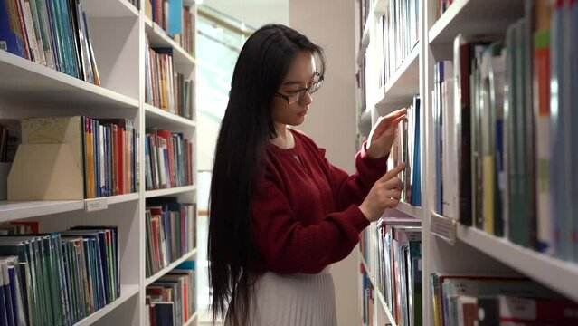 Asian student girl standing between library bookcases, young korean woman picking up book from shelf while studying in university library, using learning materials. Educational opportunities