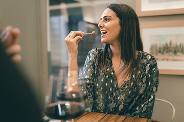 Cheerful young woman sitting at the restaurant table in front of a unrecognizable person and eating...