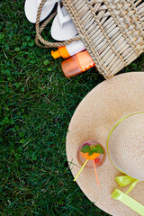 Flat lay of outdoor bright summer beach vacation or travel lifestyle concept  with straw bag, hat, flip flops, lemonade , and sunscreen spray on grass background, top view, copy space.