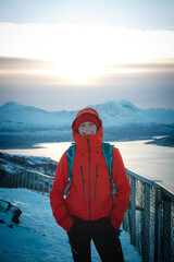 Smiling brunette hidden in a red jacket stands on top of a fjellheisen in Tromso, northern Norway during the Scandinavian winter. The joy of the climb. Candid portrait of smiling adventurer