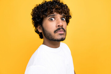 Close-up photo of a scared confused indian or arabian curly haired guy, looking at camera in disbelief, standing over isolated orange background wearing white t-shirt