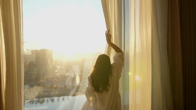 Young woman in white bathrobe opens curtains in bedroom after waking up. Lady stretching with rising hands, looking out panoramic window enjoying sunrise in early morning